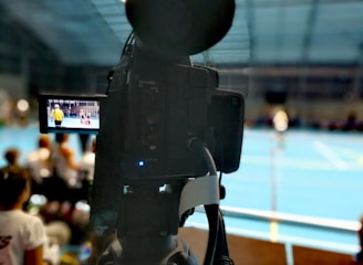 A camera on a tripod is prominently displayed with its viewfinder screen showing footage of an indoor sports event. The background is an indoor sports hall with a blue floor, and numerous spectators are faintly visible.