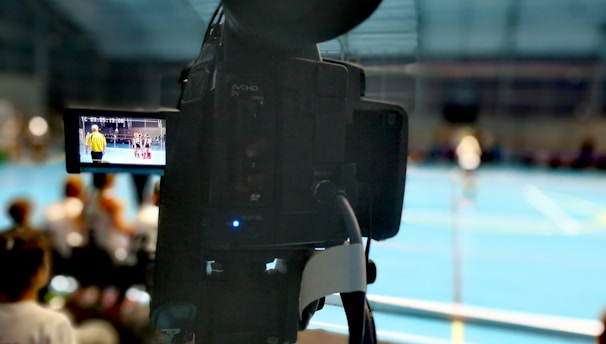 A camera on a tripod is prominently displayed with its viewfinder screen showing footage of an indoor sports event. The background is an indoor sports hall with a blue floor, and numerous spectators are faintly visible.