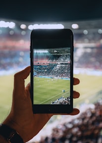A hand holding a smartphone captures a live view of a soccer stadium filled with fans and players on the field. The image on the smartphone screen is crisp, showing a wide-angle view of the game in action.