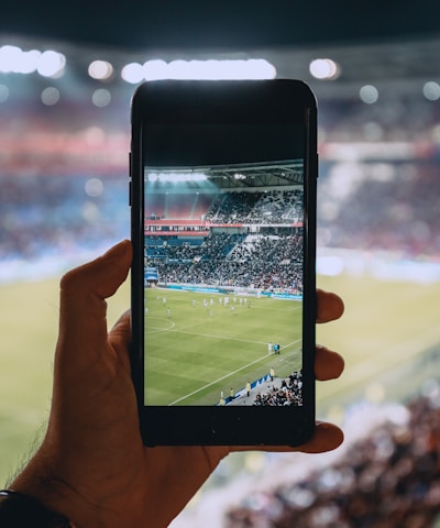 A hand holding a smartphone captures a live view of a soccer stadium filled with fans and players on the field. The image on the smartphone screen is crisp, showing a wide-angle view of the game in action.