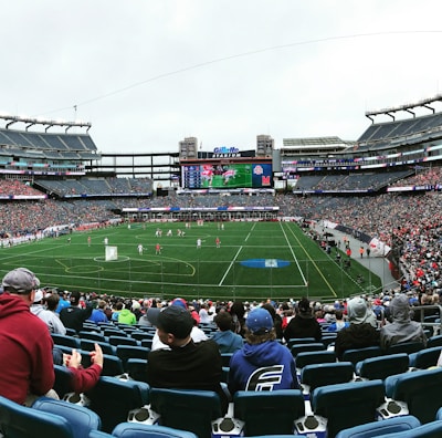A large sports stadium filled with spectators. The field is green with white markings, and players in red and white uniforms are actively engaged in a game. The stands are packed with people wearing various colored clothing, and some sections of the crowd are more densely populated. Digital screens and advertisements are visible around the stadium, adding to the vibrant atmosphere.