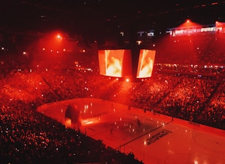 An indoor ice hockey arena filled with a large crowd, bathed in red lighting. A giant central scoreboard projects fiery visuals, illuminating the ice and audience. The atmosphere is charged, likely during a live sporting event.