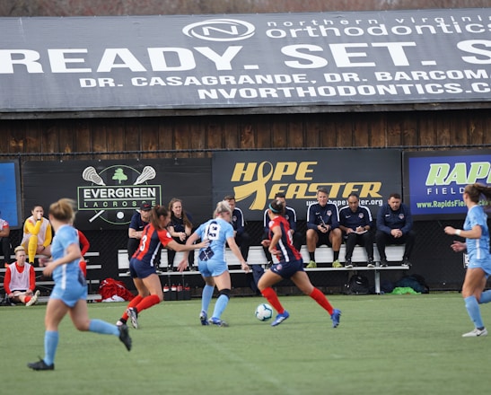 Players engage in a competitive soccer game on a field while others are seated on the bench watching attentively. The action is taking place in front of a wooden structure displaying multiple advertisements, including orthodontics, lacrosse, and field hockey.