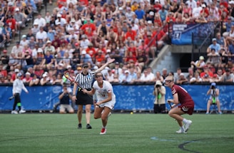 A sports event with two athletes engaged in a lacrosse game. One athlete in a white jersey is holding a lacrosse stick, appearing to prepare for a play. A referee in a striped uniform is nearby. The background features a large audience seated in a stadium, with many spectators wearing bright and casual clothing. Some photographers are also visible near the field.