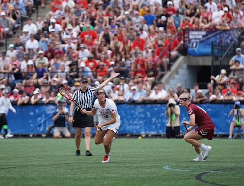 A sports event with two athletes engaged in a lacrosse game. One athlete in a white jersey is holding a lacrosse stick, appearing to prepare for a play. A referee in a striped uniform is nearby. The background features a large audience seated in a stadium, with many spectators wearing bright and casual clothing. Some photographers are also visible near the field.