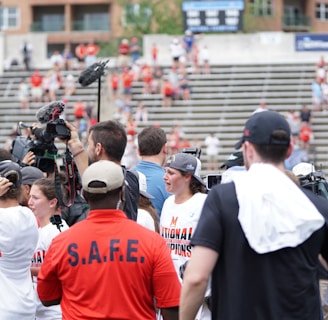A lively gathering in an outdoor sports venue with a group of people, some wearing matching shirts labeled 'National Champions.' Several individuals are holding cameras and microphones, indicating a media presence. The background shows blurred spectators sitting on bleachers.