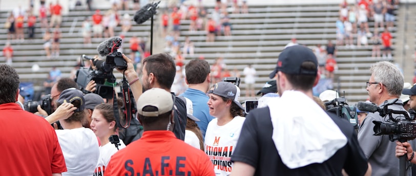 A lively gathering in an outdoor sports venue with a group of people, some wearing matching shirts labeled 'National Champions.' Several individuals are holding cameras and microphones, indicating a media presence. The background shows blurred spectators sitting on bleachers.