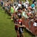 The image features a lively gathering of young soccer players and fans next to a field. Several female athletes in red and blue uniforms interact with an enthusiastic crowd standing behind a barrier. Some people in the crowd are holding objects, like pens and paper, possibly for autographs.