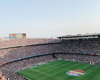 An expansive soccer stadium filled with thousands of spectators. The green field in the center features players and markings, surrounded by vibrant stands and advertising boards on the upper sides of the stadium. The audience is actively watching the game.