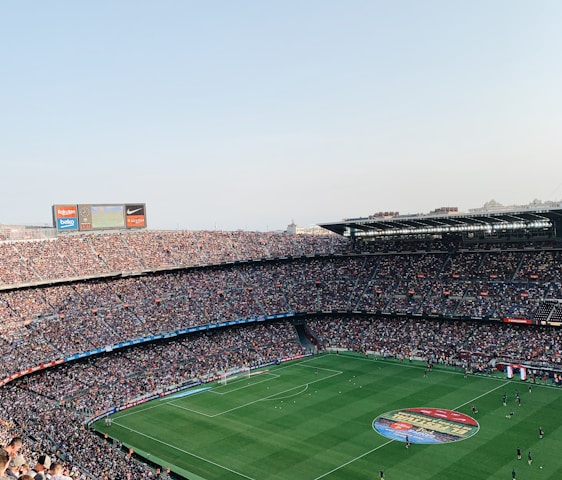 An expansive soccer stadium filled with thousands of spectators. The green field in the center features players and markings, surrounded by vibrant stands and advertising boards on the upper sides of the stadium. The audience is actively watching the game.