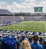 A large football stadium filled with fans watching a live game. Players are on the field, and the atmosphere is lively with a full audience. The scoreboard displays the action, and banners for the Kentucky Wildcats are visible.