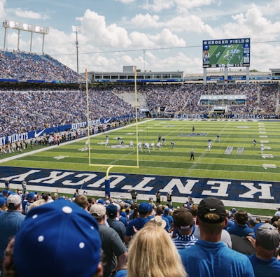 A large football stadium filled with fans watching a live game. Players are on the field, and the atmosphere is lively with a full audience. The scoreboard displays the action, and banners for the Kentucky Wildcats are visible.
