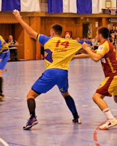 A dynamic indoor sports scene with players in motion on a court. Two athletes in blue and yellow uniforms are engaged in an intense handball game, with one player gripping the other's jersey. The background shows other players, several seated people, and advertisements on the walls.