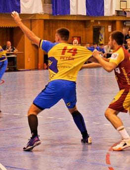 A dynamic indoor sports scene with players in motion on a court. Two athletes in blue and yellow uniforms are engaged in an intense handball game, with one player gripping the other's jersey. The background shows other players, several seated people, and advertisements on the walls.
