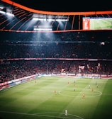 A large football stadium filled with spectators watching a game in progress. The stadium is brightly lit, with players in red and dark uniforms scattered across the green field. The scoreboard is visible, displaying game information. The atmosphere appears lively and energetic, with a sea of fans in the stands.