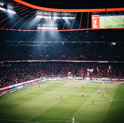A large football stadium filled with spectators watching a game in progress. The stadium is brightly lit, with players in red and dark uniforms scattered across the green field. The scoreboard is visible, displaying game information. The atmosphere appears lively and energetic, with a sea of fans in the stands.