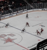 A professional ice hockey match captured from a high vantage point, featuring players in red and white uniforms moving across the rink. The scene includes a large digital screen displaying a live feed of the game and a sparsely filled audience watching from the stands.