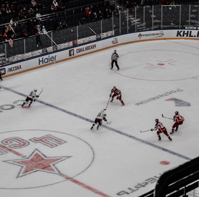 A professional ice hockey match captured from a high vantage point, featuring players in red and white uniforms moving across the rink. The scene includes a large digital screen displaying a live feed of the game and a sparsely filled audience watching from the stands.
