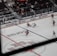 An indoor ice hockey game is taking place in an arena. The focus is on several players in action on the ice rink, with one player attempting to control the puck. The audience is visible in the stands, which are partially filled. A large overhead screen displays a live feed of the game, showing additional angles of the players. Advertising banners are positioned around the rink.