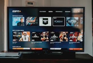 A television screen displaying the ESPN+ streaming interface with various sports-related shows and categories. The screen includes sections such as 'Browse by Shows' and 'Best of Drew Brees' with thumbnails of sports programs featuring athletes in action and analysts.