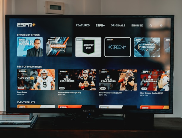 A television screen displaying the ESPN+ streaming interface with various sports-related shows and categories. The screen includes sections such as 'Browse by Shows' and 'Best of Drew Brees' with thumbnails of sports programs featuring athletes in action and analysts.