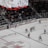 A professional ice hockey game is in progress inside an arena, with players in red and white jerseys actively engaging on the rink. The scoreboard above displays the score and game details, while spectators are seated in the stands. A large screen shows a live feed of the match action.