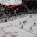 A professional ice hockey game is in progress inside an arena, with players in red and white jerseys actively engaging on the rink. The scoreboard above displays the score and game details, while spectators are seated in the stands. A large screen shows a live feed of the match action.