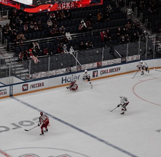 An ice hockey game in progress at a large indoor arena, with players in action on the rink. Spectators are seated in the stands, and a large digital screen displays the game above the action.