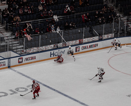 An ice hockey game in progress at a large indoor arena, with players in action on the rink. Spectators are seated in the stands, and a large digital screen displays the game above the action.