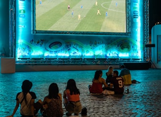 A large outdoor screen displays a soccer match at night. A small group of people sit on a cobblestone surface, watching the game. The screen is surrounded by a bright blue glow, creating a lively atmosphere.