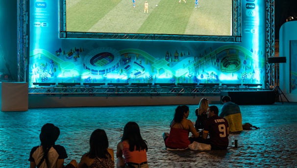 A large outdoor screen displays a soccer match at night. A small group of people sit on a cobblestone surface, watching the game. The screen is surrounded by a bright blue glow, creating a lively atmosphere.