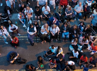 A large group of people gathered outdoors, watching a screen that is displaying a man. The crowd is diverse in age and appears interested and engaged, with many sitting closely together, some standing. Many are dressed casually, some in sports attire, and a few waving or holding flags. The event seems to have a communal, festive atmosphere.