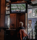 A person with short, light-colored hair is sitting in a cozy, wood-paneled interior of what appears to be a bar or cafe, watching a football game on a mounted TV screen. A warm-colored jacket is draped over their chair. The TV displays a live football match with a clear scoreboard and players on the field. The space is decorated with string lights, and an American flag is visible outside through a window next to some greenery.