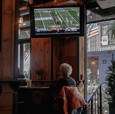 A person with short, light-colored hair is sitting in a cozy, wood-paneled interior of what appears to be a bar or cafe, watching a football game on a mounted TV screen. A warm-colored jacket is draped over their chair. The TV displays a live football match with a clear scoreboard and players on the field. The space is decorated with string lights, and an American flag is visible outside through a window next to some greenery.
