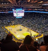 An indoor basketball arena filled with spectators. The court features a central logo and is surrounded by partially occupied tiered seating. A large scoreboard hangs above the center of the court, displaying the game score and time. Ambient lighting highlights the players on the court and the vibrant atmosphere of a live sports event.