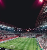 A large, illuminated stadium packed with spectators, showcasing a vibrant soccer match. The field is green with visible players in red and white uniforms, and the stands are filled with red and white-clad fans. The stadium structure is visible above, with bright lights casting a glow.