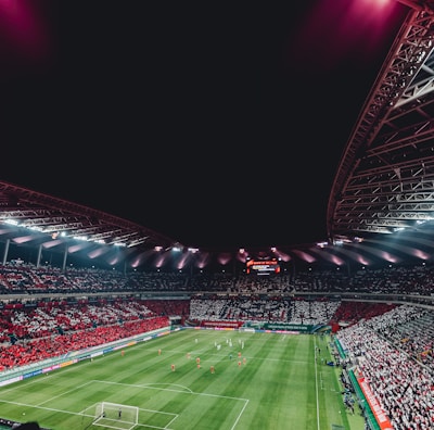 A large, illuminated stadium packed with spectators, showcasing a vibrant soccer match. The field is green with visible players in red and white uniforms, and the stands are filled with red and white-clad fans. The stadium structure is visible above, with bright lights casting a glow.