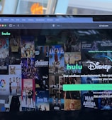 A laptop screen displays a web page featuring a mix of movie and TV show covers, highlighting popular titles and a Hulu and Disney+ bundle promotion. There are visible icons and images of films and shows alongside promotional text offering entertainment and live sports.