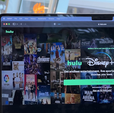 A laptop screen displays a web page featuring a mix of movie and TV show covers, highlighting popular titles and a Hulu and Disney+ bundle promotion. There are visible icons and images of films and shows alongside promotional text offering entertainment and live sports.
