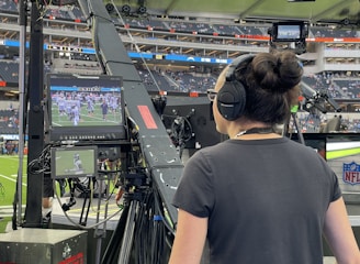A person with headphones is operating a large camera setup at a sports stadium. The camera equipment is focused on a screen showing a live sports event taking place on a football field. The stadium is filled with spectators, and the atmosphere is busy with technical setup and monitors.