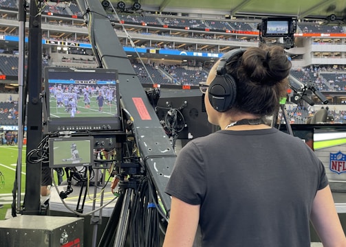 A person with headphones is operating a large camera setup at a sports stadium. The camera equipment is focused on a screen showing a live sports event taking place on a football field. The stadium is filled with spectators, and the atmosphere is busy with technical setup and monitors.
