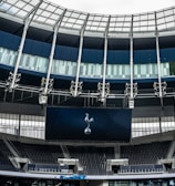 A modern stadium interior featuring a large digital screen with a logo prominently displayed. The seating areas are empty, with dark-colored seats and some structural elements like beams and glass walls visible. A combination of dark blue and neutral tones are prominent.