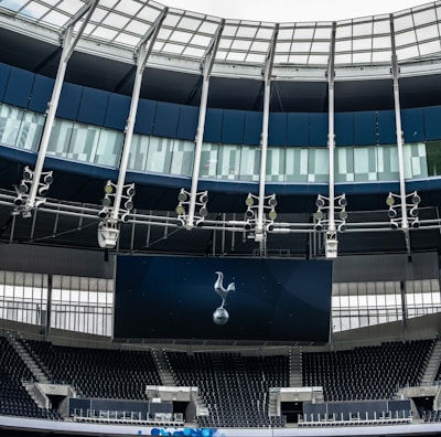 A modern stadium interior featuring a large digital screen with a logo prominently displayed. The seating areas are empty, with dark-colored seats and some structural elements like beams and glass walls visible. A combination of dark blue and neutral tones are prominent.